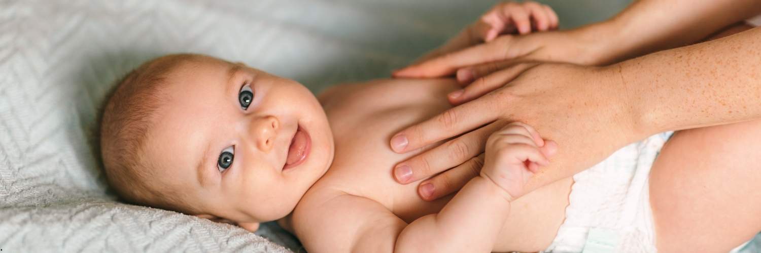 Un bébé allongé sur une table à langer avec sa maman en train de lui passer de la crème sur le corps