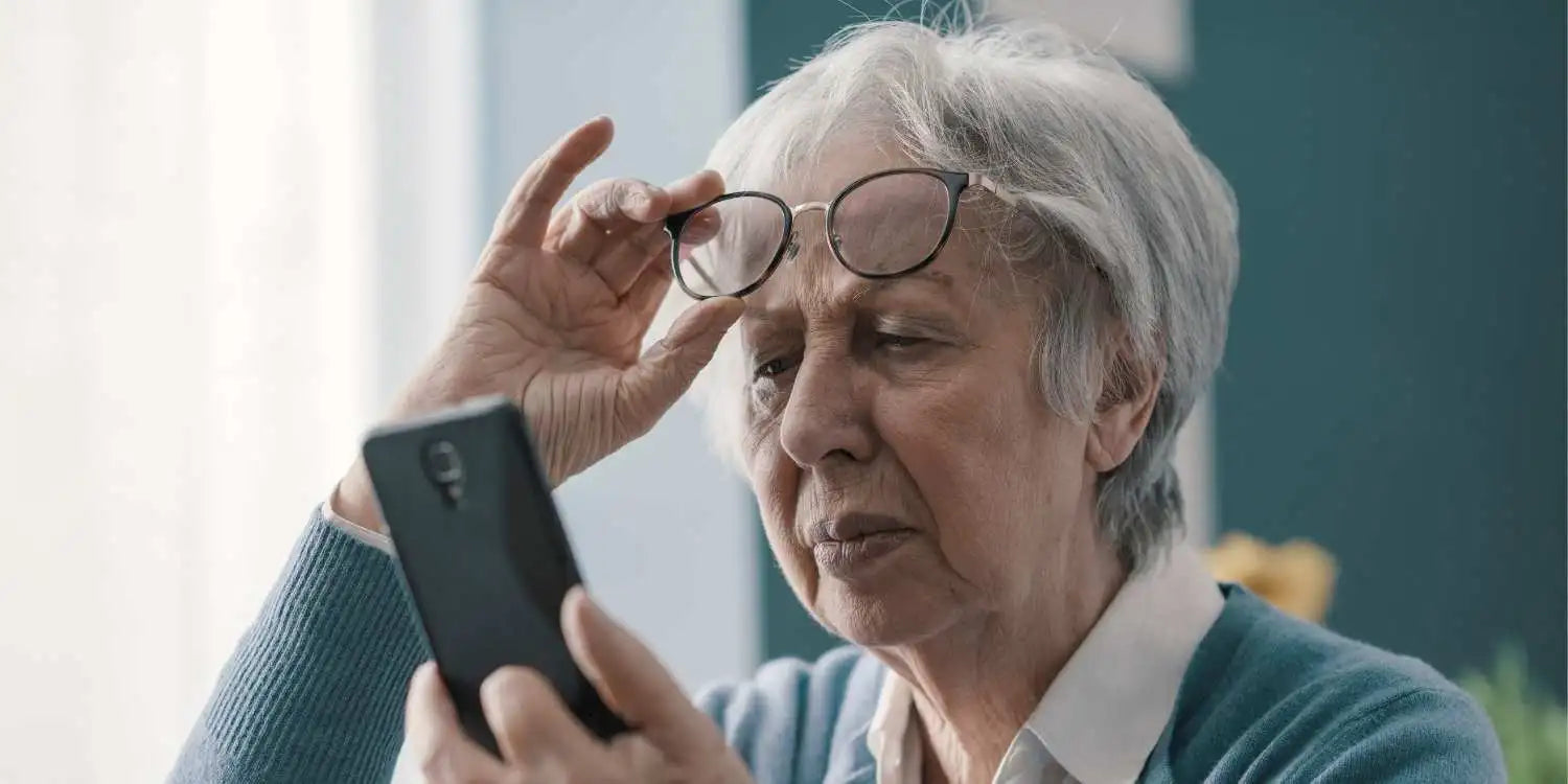 Une vieille dame soulevant ses lunettes ayant visiblement du mal à lire le contenu de l'écran de son téléphone