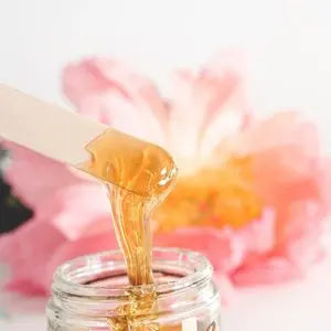Cire d'épilation au miel coulant d'une spatule en bois devant une pivoine rose