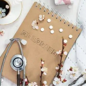 Carnet en spirale avec le mot 'Ménopause' écrit dessus, entouré de branches fleuries, de pilules blanches et d'un stéthoscope, posé sur une surface claire avec une tasse de café et un tissu blanc.