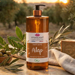 Un savon liquide d'alep de 1L dans un décor méditarénéen avec des vignes en arrière plan et un sachet d'olive à côté du produit.