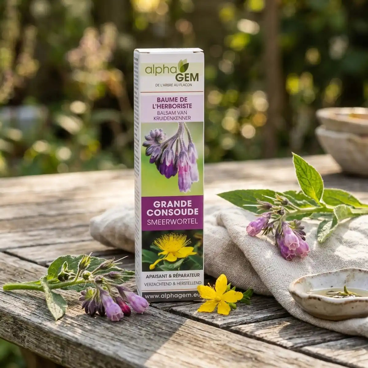 Baume à la Grande Consoude Bio Alphagem 50g sur table en bois, entouré de fleurs de consoude et de millepertuis, lumière naturelle