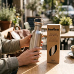Bouteille en acier inoxydable 500ml Bambaw au bouchon bambou, posée sur une table en bois ensoleillée à côté de son étui en carton kraft