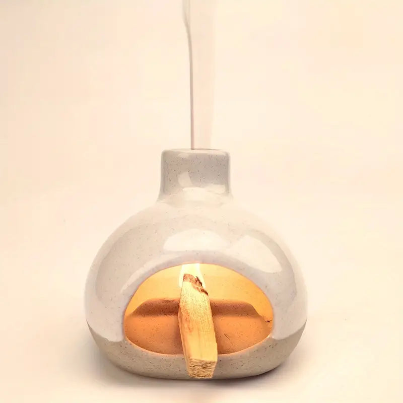 Un brûleur d'encens en céramique bicolore avec collecteur de cendres, en fonctionnement, avec du palo santo en train de bruler et de la fumée s'échappent de la cheminée