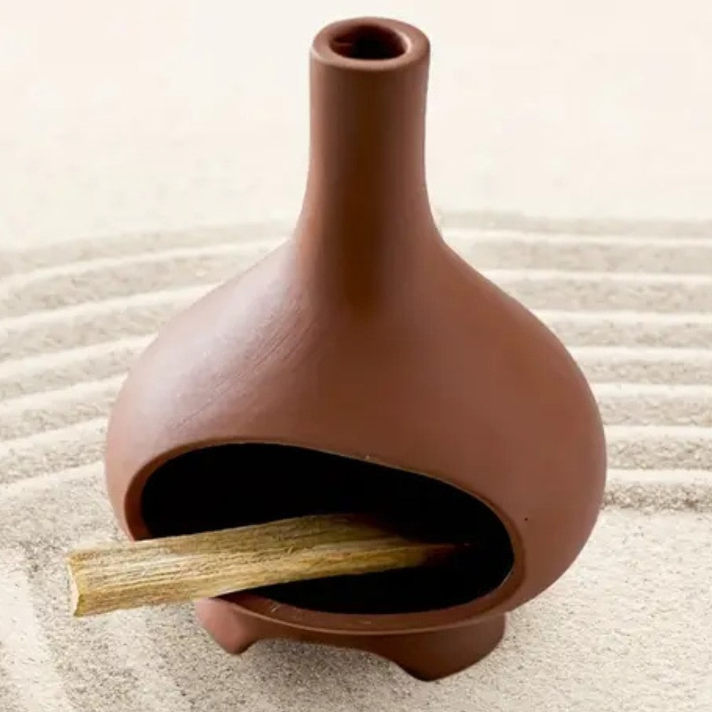 Un brûleur d'encens pour Palo Santo en céramique couleur terre cuite posé sur un lit de sable blanc, avec du palo santo en train de bruler