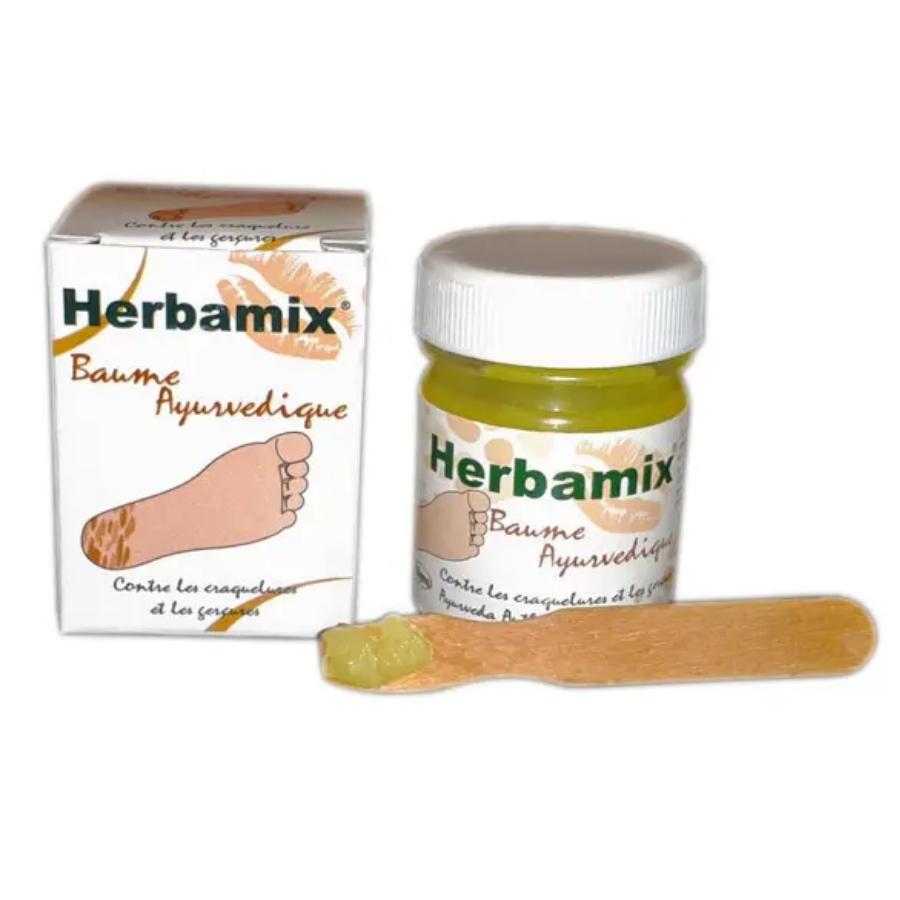Un Baume Ayurvédique Herbamix pour les Pieds, Mains & Lèvres avec son emballage carton et une spatule en bois contenant un peu de baume sur le bout