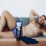 Un tube de Crème dépilatoire zones intimes Blondépil Homme posé sur une table avec un homme en sous-vêtements à l'arrière plan