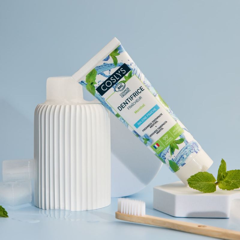 Un dentifrice fraicheur menthol de 75 ml de la marque Coslys posé devant une brosse à dent et des feuilles de menthe