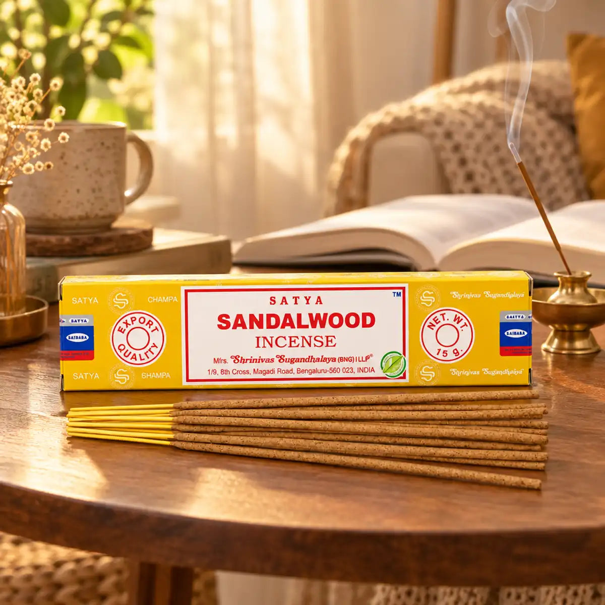 Boîte jaune et bâtonnets d'encens Satya Bois de Santal 15g, posés sur une table dans un coin lecture chaleureux avec porte-encens en laiton