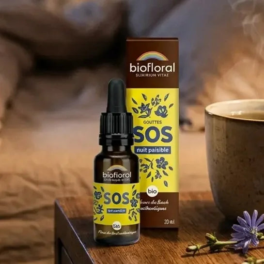 Flacon compte-gouttes SOS Nuit Fleurs de Bach Biofloral 20 ml sur une table de chevet en bois, à côté d'une tasse de tisane et de fleurs sauvages bleues