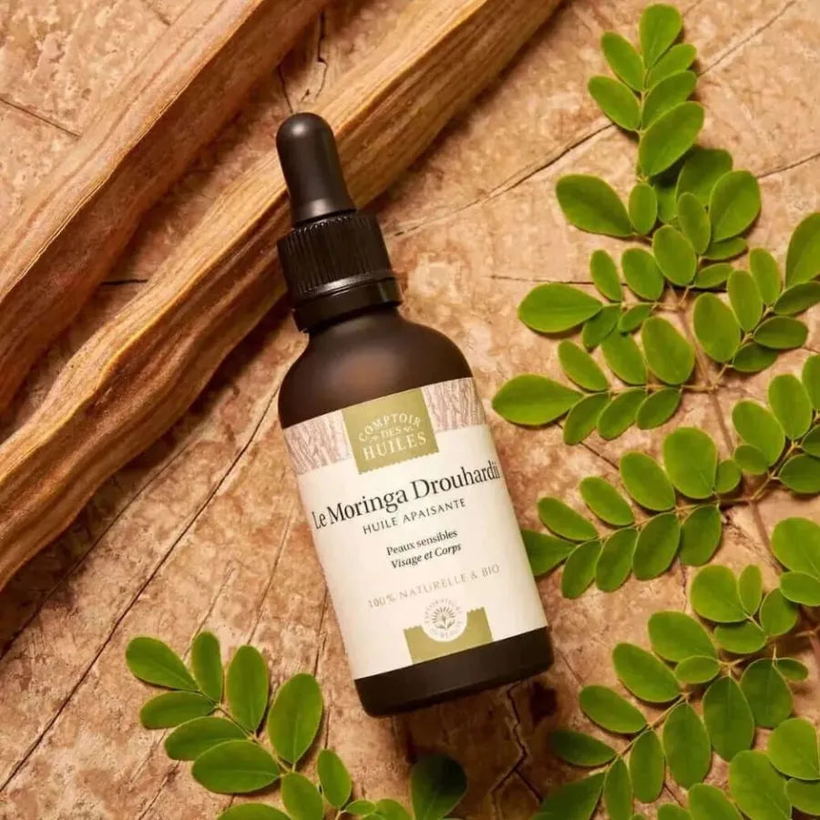 Une huile de moringa Drouhardii de la marque comptoir des huiles en 50 ml posée sur un tronc d'arbre avec des feuilles de moringa