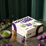 Un coffret de match et d'ube avec un fouet, un bol de poudre de matcha et de l'ube avec des boules de noël, des pommes de pins et de l'eucalyptus