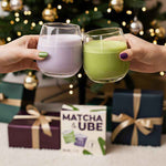 deux mains tchinant avec un verre d'ube et de matcha devant un sapin de noël