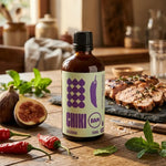 Sauce piquante bio Calabria Chiki Bam 100 mL, flacon ambré sur table rustique avec piments frais, figue et menthe