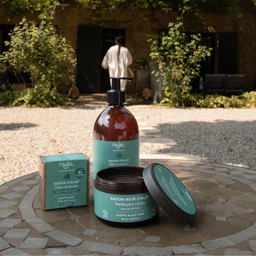 Savon d'Alep liquide Najel 500ml avec d'autres produits de la même gamme posés sur une table en extérieur