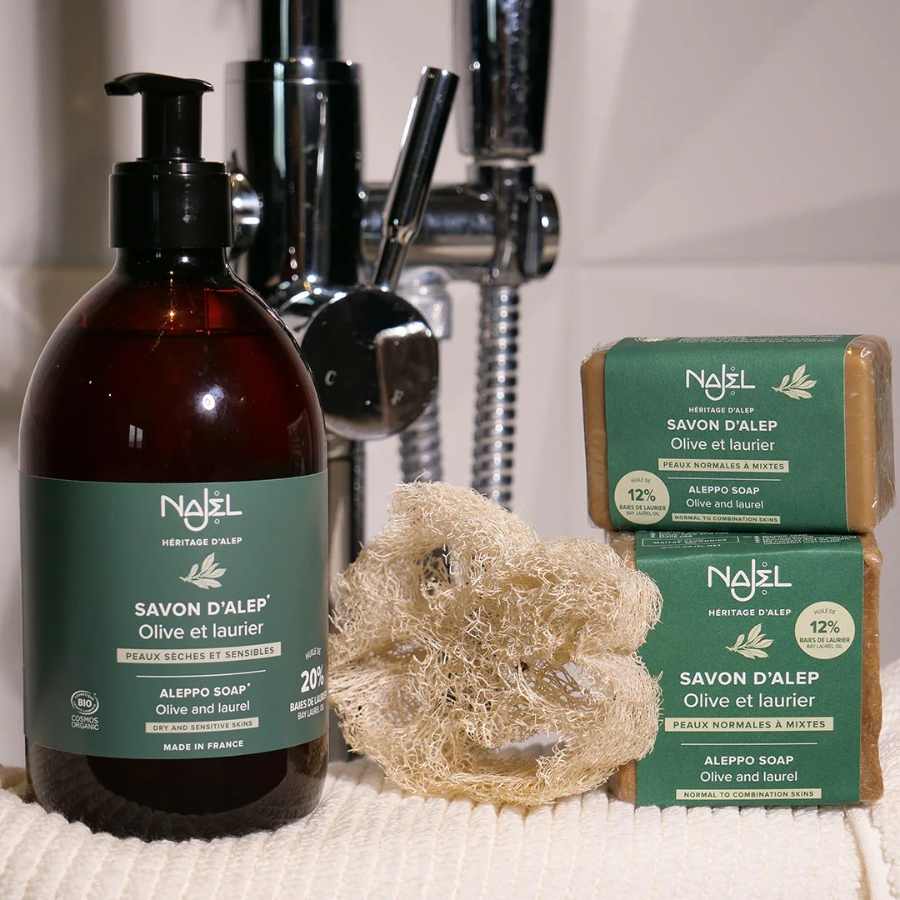 Savon d'Alep liquide 20% Najel 500ml en flacon dans une salle de bain à côté d'une éponge et de savons d'alep solides
