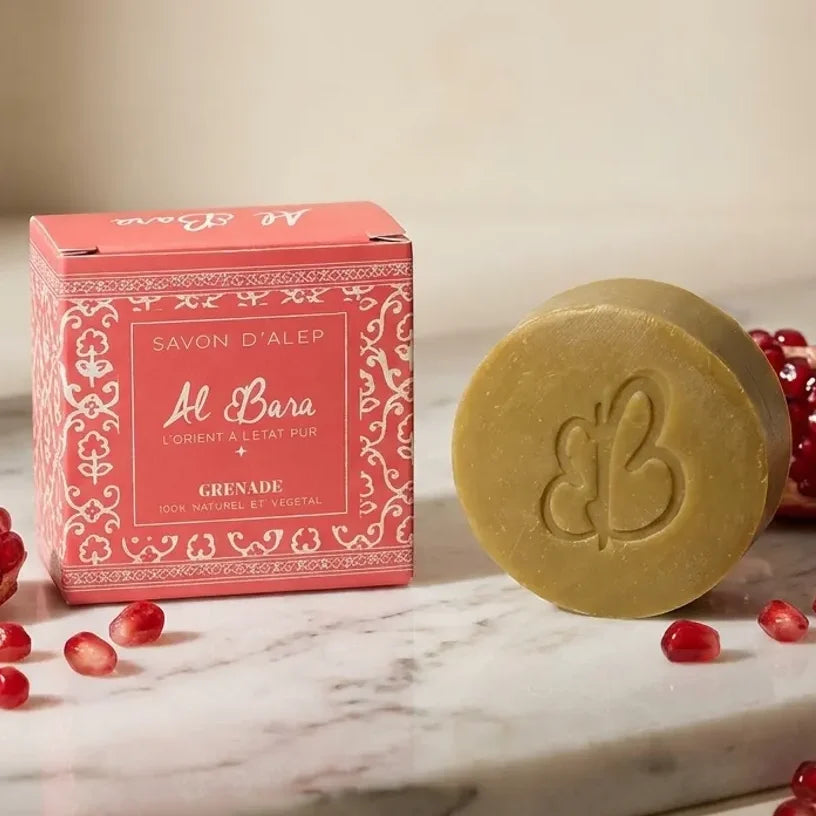 Savon d'Alep parfumé à la grenade 50g Al Bara, pain de savon doré à côté de son coffret rose orné de motifs orientaux, présenté sur un marbre avec des grains de grenade