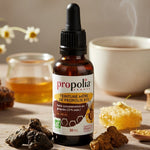 Flacon de Teinture Mère de Propolis Bio Propolia au premier plan, avec un cadre d'éléments naturels sur une table en bois clair. On y voit des morceaux de propolis brute, des rayons de miel, des fleurs de camomille, un pot de miel liquide et une tasse de tisane fumante. L'ensemble évoque la santé, le bien-être et la nature.