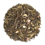 Sachet de thé vert bio « Caprice Gourmand » de la marque Les Jardins de Gaïa, aromatisé à la rhubarbe et à la fraise, présenté à côté d'un tas de feuilles de thé en vrac parsemé de morceaux de fruits.